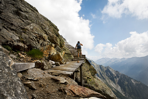 Young man with backpack hiking in scenic mountain landscape of the Dolomite alps in Italy. Recreational pursuit under a cloudy sky. Natural landmark. Walking up a steep, wooden path attached to the rock in the nature reserve Rieserferner-Ahrn near Antholz. XXXL (Canon Eos 1Ds Mark III)