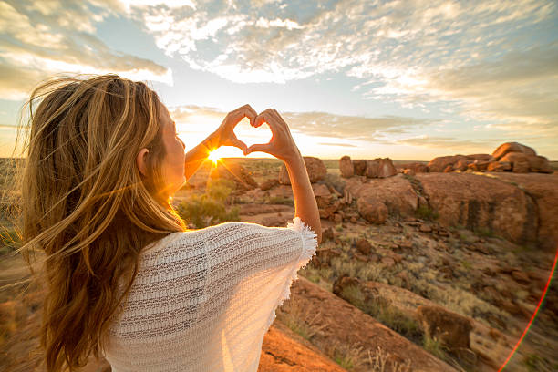 若い女性は壮大な風景の日の出にハートシェイプの指のフレームを作ります - devils marbles ストックフォトと画像