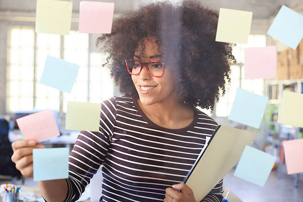 Smiling young woman taking colorful reminders from glass wall Smiling young woman with red eyeglasses holding documents and taking sticky notes from glass wall. Focus on foreground. workplace absence stock pictures, royalty-free photos & images