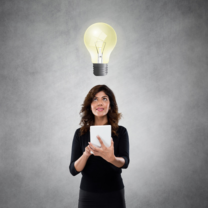 Young businesswoman standing working on digital tablet with glowing light bulb over her head as symbol aspirations.