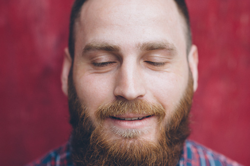 bearded young man on a background of a red wall dreaming with closed eyes