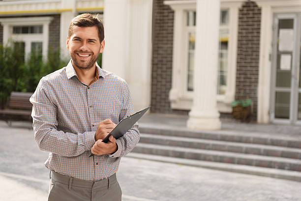Smiling Real Estate Agent standing outside modern house Real professional. Portrait of male Real Estate Agent standing outside residential property, holding clipboard and smiling man agent stock pictures, royalty-free photos & images