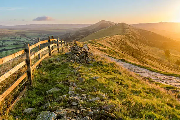 A fresh Autumnal sunrise a long The Great Ridge in the English Peak District. The image features a stone path that runs a long the ridge with a fence, bathed in vibrant golden light from the rising sun.