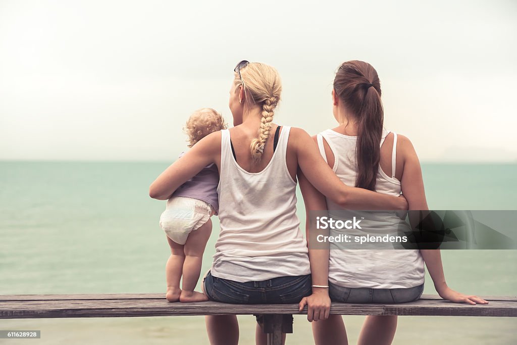 Loving mother embracing her children looking into the distance Loving mother embracing her children sitting on wooden bench at tropical beach during vacation. They together looking into the distance. Concept for togetherness and  bright future Lesbian Stock Photo