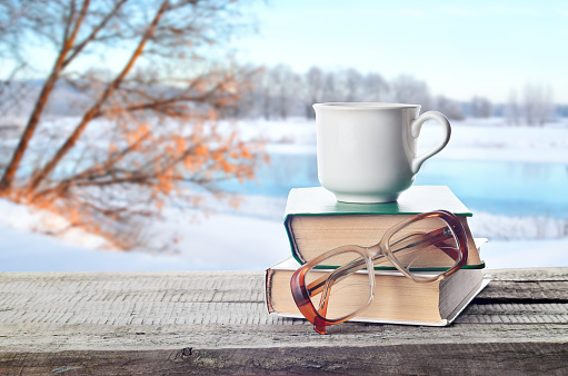 Hot coffee or tea, cocoa, chocolate cup on book and eyeglasses outdoors in winter. Pile of books, glasses and cup in nature.