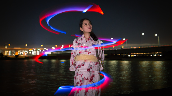 Young Yukata woman standing nearby river with red blue light