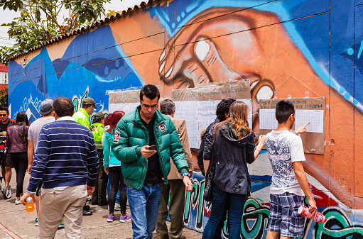 Bogota, Colombia - October 02, 2016: Voters check lists posted on the wall of a polling station on Carrera 9, in the Andean capital city of Bogota, in the South American country of Colombia, to find out the exact table number where they can vote on the historic Peace Referendum. The lists are hung on a wall that has a mural painted in the bright colours of Latin America, dedicated to the Peace Process. Photo shot in the afternoon sunlight.