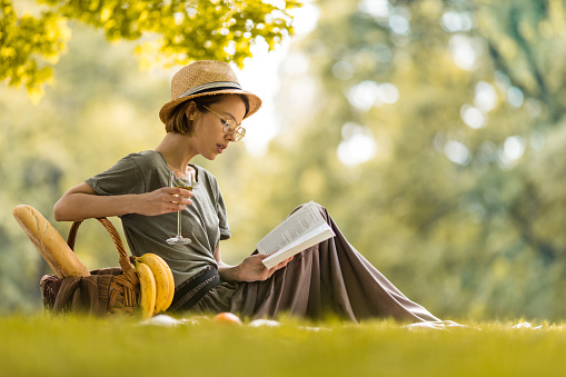 Relaxed woman enjoying in nature at springtime and reading a book while drinking white wine.