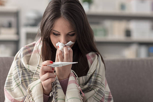 Sick woman taking temperature Sick young woman at home on the sofa, she is covering with a blanket, taking temperature and blowing her nose with a tissue fever stock pictures, royalty-free photos & images