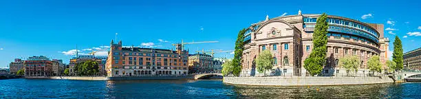 Tourists and locals enjoying the summer sunshine on Helgeandsholmen in the historic Gamla Stan old town of Stockholm, overlooked by the Riksdagsjuset parliament buildings in the heart of Sweden's vibrant capital city.