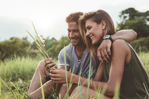 Shot of smiling young couple sitting together outdoors. Man and woman relaxing in the grass field holding wild flower.