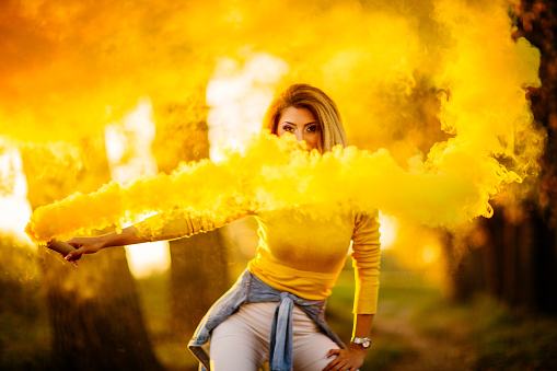 Young pretty girl outdors in a park, on a lovely autumn day, holding a torch, there is yellow smoke all around her