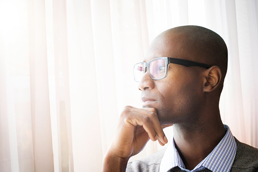 Pensive mature bald black man by a window resting his chin on his hand. He is wearing glasses, a shirt and a sweater.