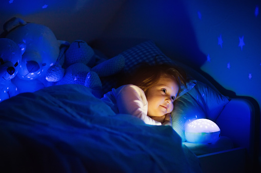 Little girl reading a book in bed. Dark bedroom with night light projecting stars on room ceiling. Kids nursery and bedding. Children read before bedtime. Toddler child playing with lamp and bear toy.