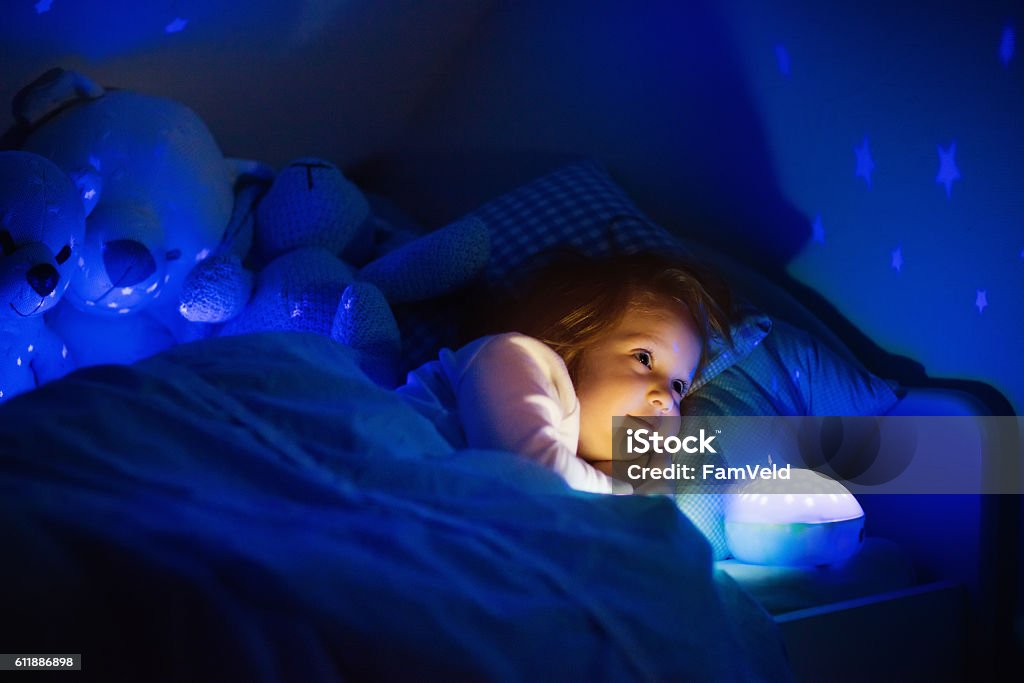 Little girl in bed with night lamp in dark nursery Little girl reading a book in bed. Dark bedroom with night light projecting stars on room ceiling. Kids nursery and bedding. Children read before bedtime. Toddler child playing with lamp and bear toy. Child Stock Photo