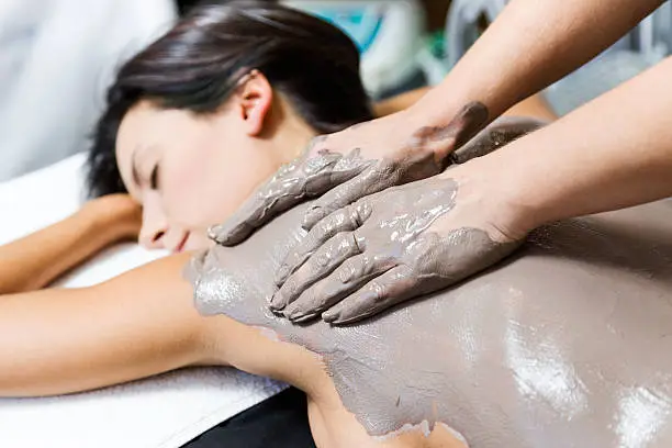 Portrait of beautiful young woman having clay body mask apply by beautician. Detox ritual.