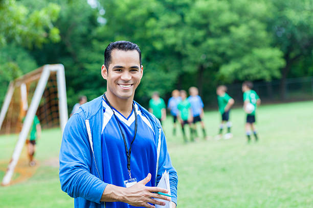Handsome mid adult Hispanic soccer coach smiles confidently on the sidelines during soccer game. He is holding a clipboard and is wering a blue jersey with blue hoodie. He is looking at the camera. His team and the opposing game are on the playing field along with the goal. Trees line the playing field.