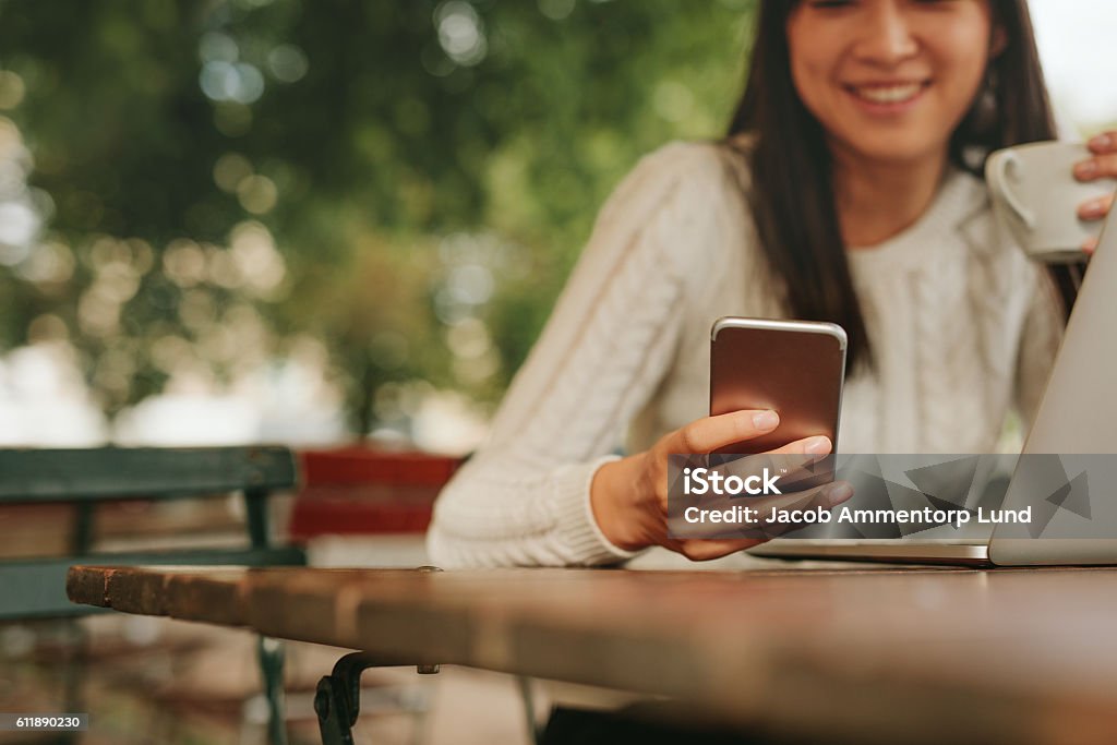 Young woman in a cafe using mobile phone Young woman in a cafe reading a text message from her mobile phone. Chinese female sitting at cafe table with laptop and using smart phone. Mobile Phone Stock Photo