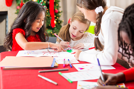 A group of diverse elementary age girls sitting at a table and making Christmas greeting cards and crafts together. The girls are happy and talking to one another as they decorate their cards in the living room.