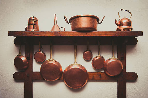 Different kind of vintage copper cookware, pans, coffee pot and funnel hanging on wooden shelf in kitchen with white rough wall