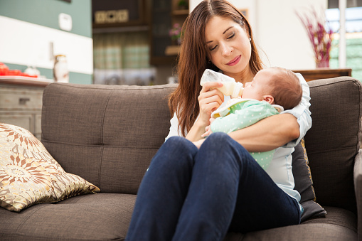 Good looking young woman sitting at home with her newborn baby and feeding her milk from a bottle
