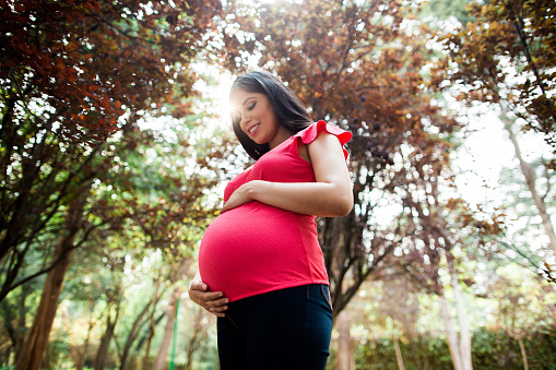 A latin pregnant woman standing, holding belly and looking at it in a horizontal medium shot outdoors.