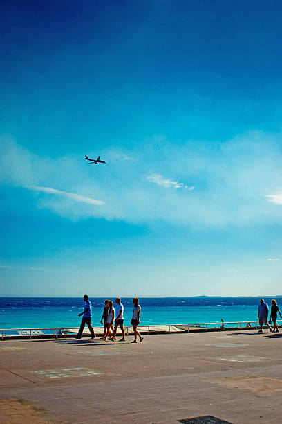 Nice, France - July 29, 2015: Family walking along the promenade in front of the beach in Nice, France. This has a polarized blue sky with a passenger airliner flying overhead.