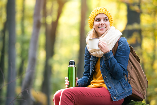 Beautiful young woman on a hike in autumn forest, having tea or coffee in a cup from thermos