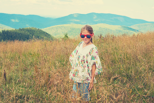 Little girl in hippie style posing on a mountain meadow. Bright red sunglasses and colorful blouse. Vintage. Toned image
