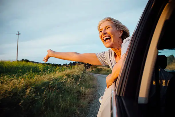 Mature woman leaning out the window of her car having fun as the sunset over the Tuscany Landscape. She smiles and her arm is outstretched.