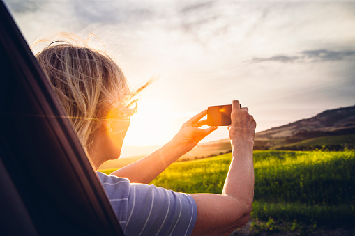 Mature woman taking a picture leaning out the window of her car of the sunset over the Tuscany Landscape. Lens flare as she uses her smart phone.