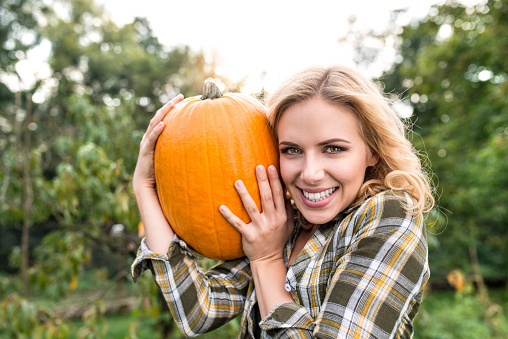 Beautiful young blond woman in checked shirt working in her garden harvesting pumpkins. Autumn nature.