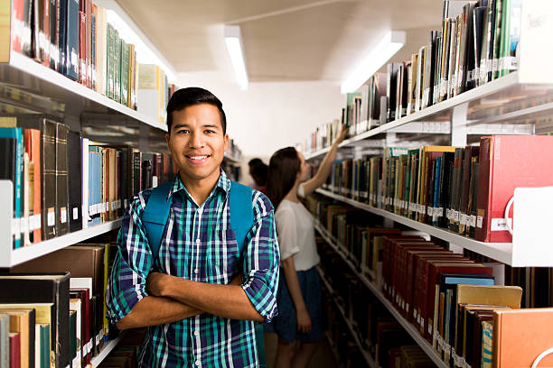 istock Latin student standing between bookshelves with arms crossed 612248644