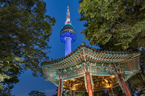 Beautiful illuminated N Seoul Tower between trees with colorful Golden Pavilion in the front at twilight - night in Namsan Park on Namsan Mountain, Seoul, South Korea, Asia. 