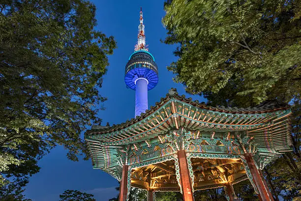 Beautiful illuminated N Seoul Tower between trees with colorful Golden Pavilion in the front at twilight - night in Namsan Park on Namsan Mountain, Seoul, South Korea, Asia. 