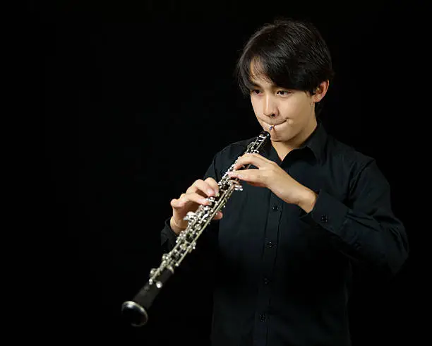 Waist up shot of a boy playing an oboe. This is a studio shot on a black background. He is formally dressed wearing a plain black shirt.