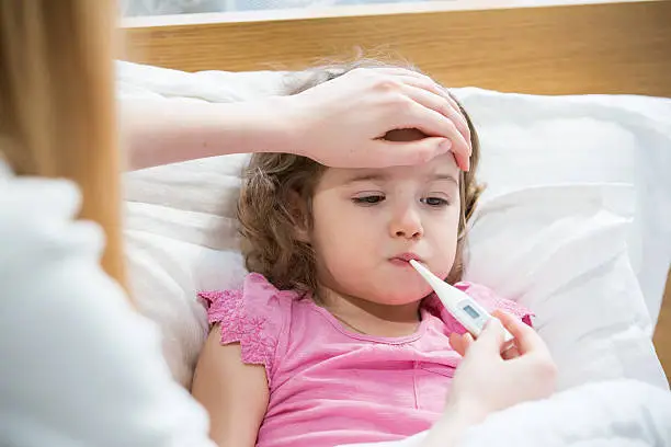 Mother measuring temperature of her ill kid. Sick child with high fever laying in bed and mother holding thermometer. Hand on forehead.