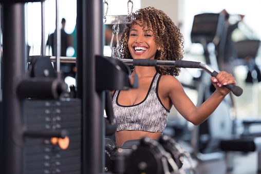 Portrait of happy athlete woman training on lateral pull-down weights exercise machine. Woman holding wide grip bar, looking at camera with a friendly smile.