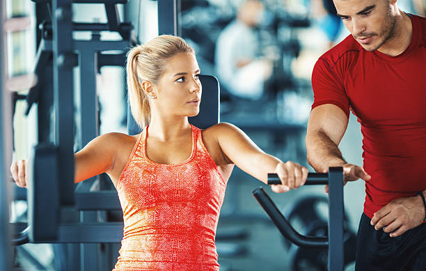 Woman exercising in a gym with an instructor. Closeup front view of mid 20's blond woman working out on a chest press machine. Her instructor is standing next to her and making sure she's doing it properly. blonde female bodybuilders stock pictures, royalty-free photos & images