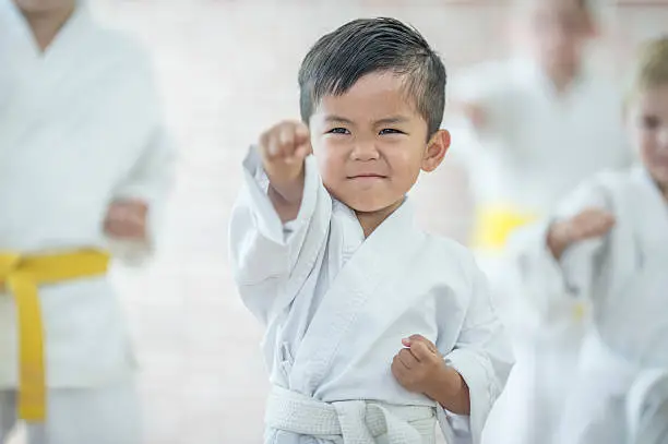 A multi-ethnic group of elementary age children are taking a karate class together at a health club. A cute little boy is raising his fist in the air to punch.