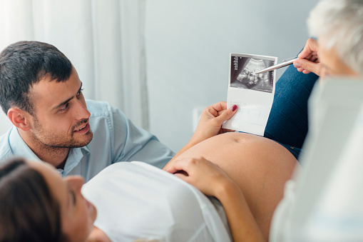 Pregnant woman and her female doctor in a consultation. Woman lying and holding ultrasound image and female doctor pointing and talking. Young woman husband is beside her and they are listening doctor.