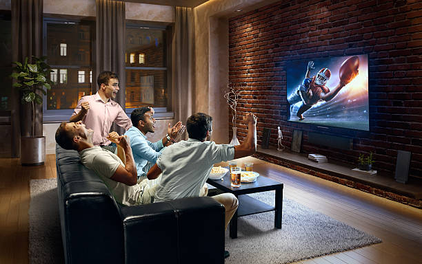 Young men cheering and watching American football game on TV A group of young male friends cheering and watching American football game on TV. They are sitting on a sofa in the modern living room. The TV set is on the loft brick wall. It is evening ouside the window. keep an eye on stock pictures, royalty-free photos & images