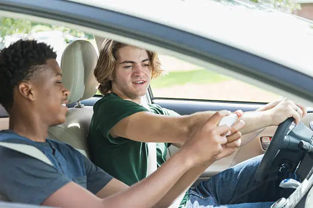 An 18 year old teenage boy or young man sitting in the driver's seat of a car. His African American passenger is showing him something on his mobile phone.