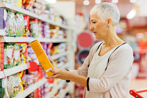 Mature Woman Groceries Shopping. Mature woman shopping in local supermarket. Standing by produce stand and choosing cereals. Holding box and reading nutrition label. breakfast cereal photos stock pictures, royalty-free photos & images