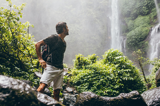 Young man with backpack standing near a waterfall in forest. Male hiker in the nature during rain.