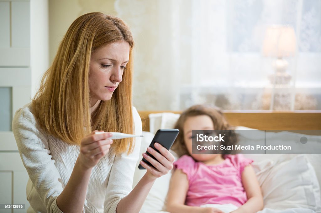 Sick child with high fever Mother measuring temperature of her ill kid. Sick child with high fever laying in bed and mother holding thermometer. Mother with cell phone calling to doctor Illness Stock Photo