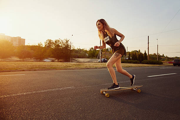 hermosa joven con tatuajes montando longboard en clima soleado - patinaje en tabla larga fotografías e imágenes de stock