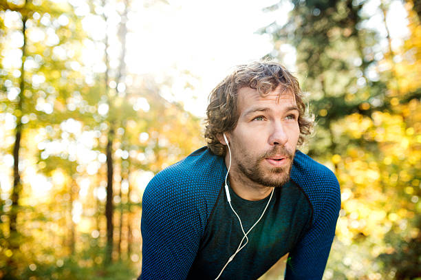 Young handsome runner with earphones outside in autumn nature Young handsome runner with earphones in his ears, listening music, outside in sunny autumn nature, resting, breathing out exhaling stock pictures, royalty-free photos & images