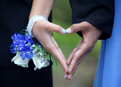 A couple going to the prom are forming a heart shape with their hands.  She is wearing a blue and white wrist corsage