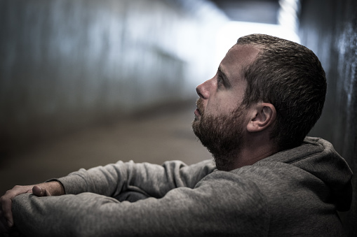 Close up image of a young adult male of caucasian ethnicity sitting in a subway tunnel and begging for money. He looks sad and depressed, and his face is bearded. The image is desaturated to add to the melancholy feel. Horizontal image with copy space.